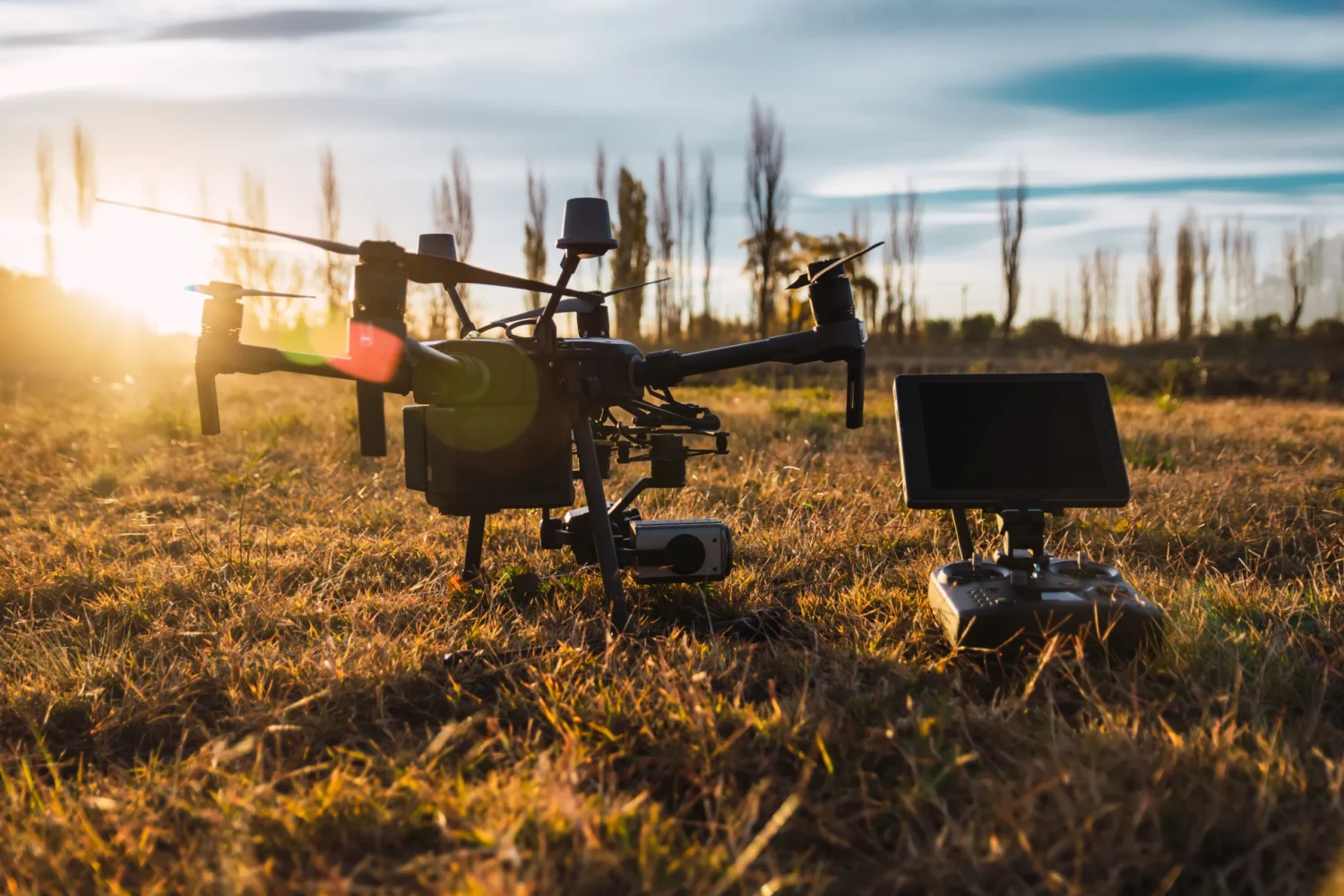 A drone and its controller sit on grassy terrain at sunset, with tall trees silhouetted in the background.